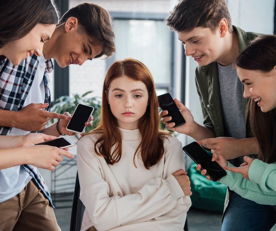 Młoda, smutna dziewczyna, rude włosy, otoczona przez nastolatków z telefonami, wyśmiewających ją